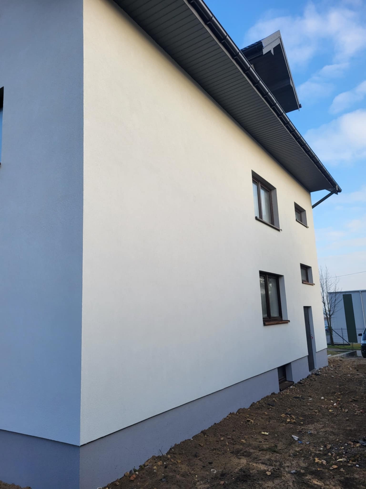 Boczny widok domu po malowaniu elewacji na biało z szarym cokołem i dachem. Widoczne okna z brązowymi ramami i fragment nieba.