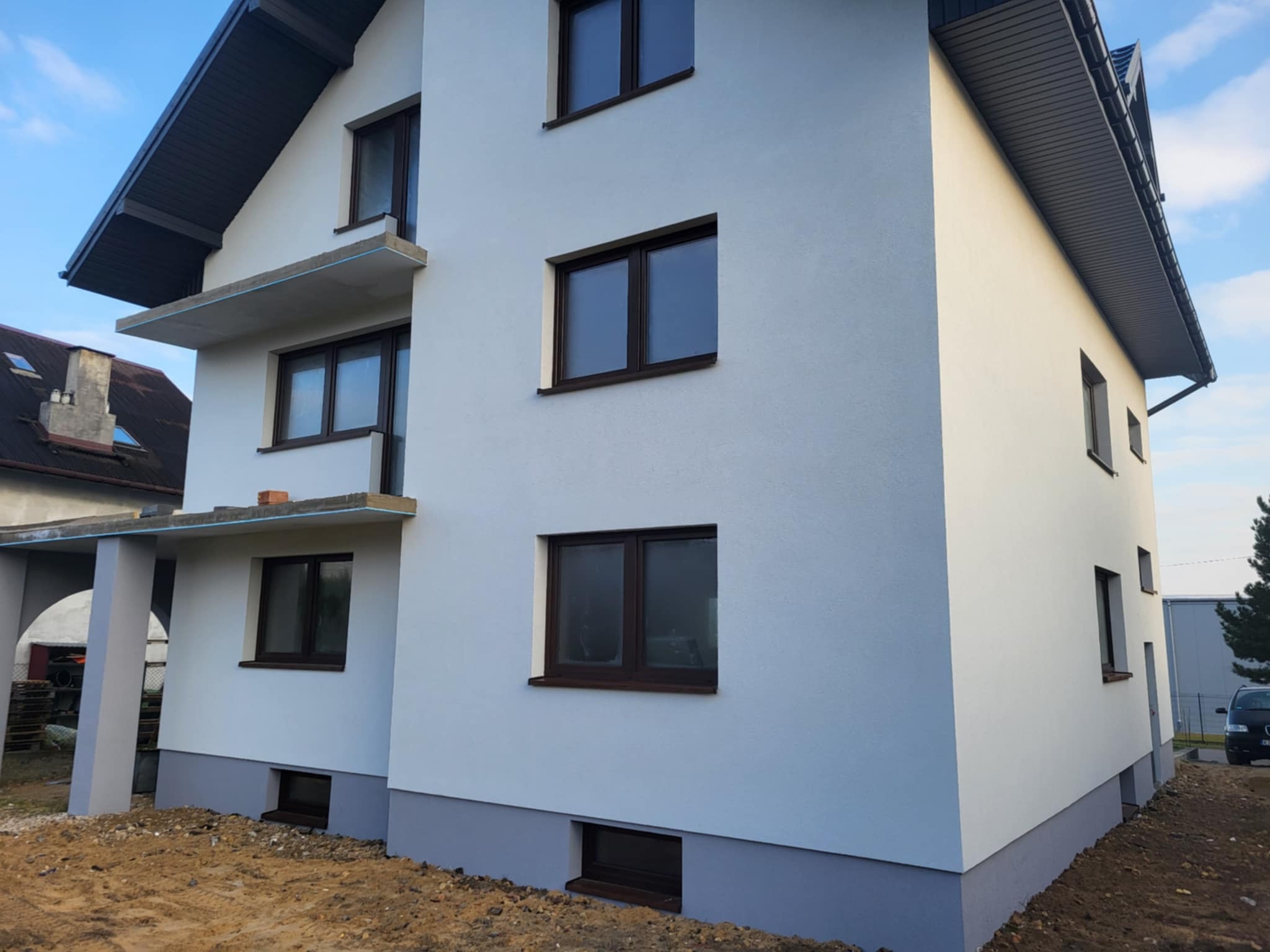 Biała elewacja dwukondygnacyjnego domu z ciemnymi oknami i szarą podmurówką, widok z boku, częściowo widoczne rusztowanie balkonu, błękitne niebo w tle.