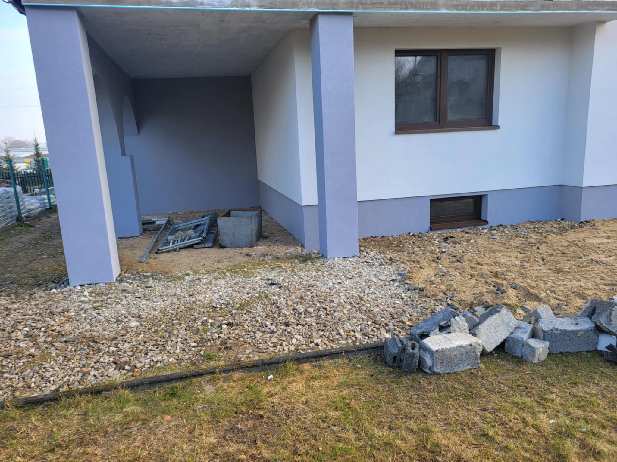Świeżo otynkowana elewacja domu jednorodzinnego z szarą podmurówką i kolumnami. Na pierwszym planie gruz i kamienie, sugerujące prace wykończeniowe.