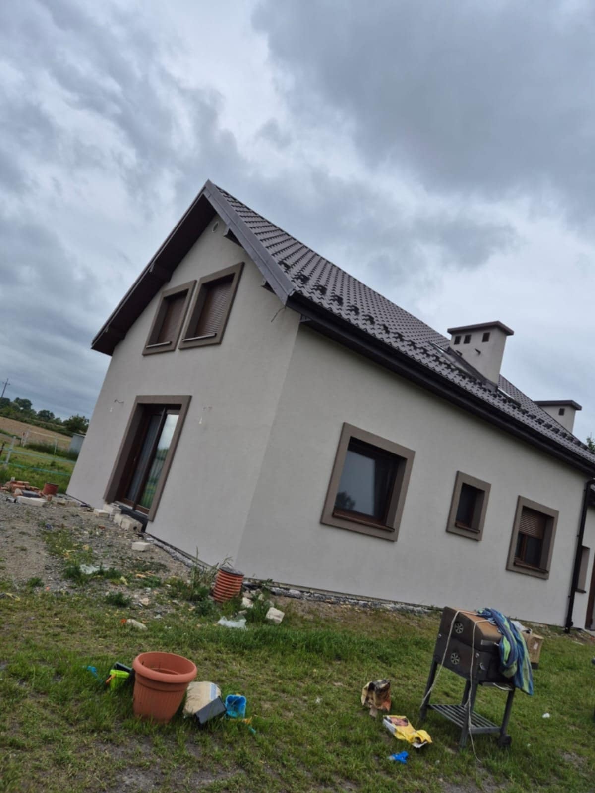 Dom jednorodzinny z beżową elewacją i brązowymi ramami okiennymi, widoczny z boku na tle pochmurnego nieba. Wokół domu trawa i elementy budowlane.