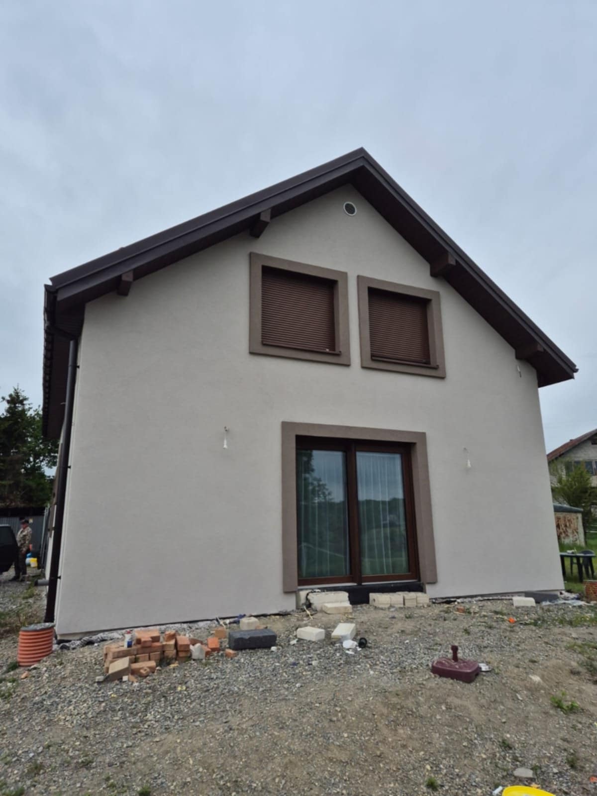 Dwukondygnacyjny dom z jasną elewacją i brązowymi ramami okiennymi, widoczny z dołu, z cegłami i gruzem u podstawy, sugerujący prace wykończeniowe.