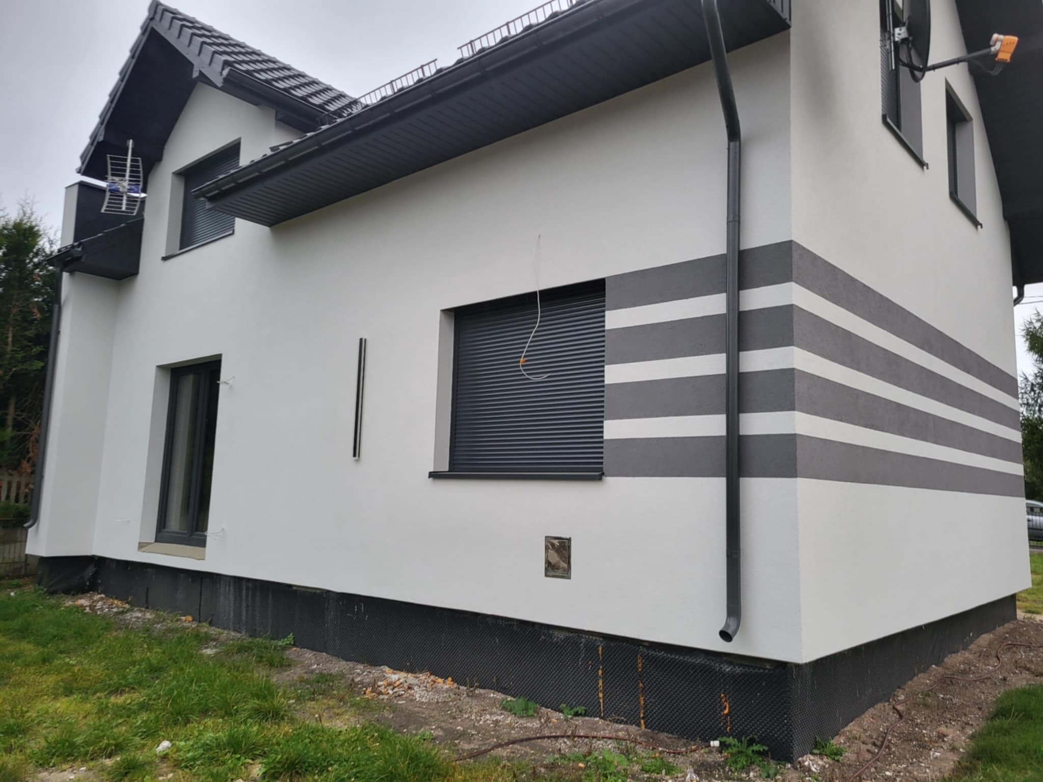 Ocieplony dom jednorodzinny z szarymi pasami na elewacji, czarnymi ramami okiennymi i orynnowaniem. Widoczna izolacja fundamentów i antena satelitarna.