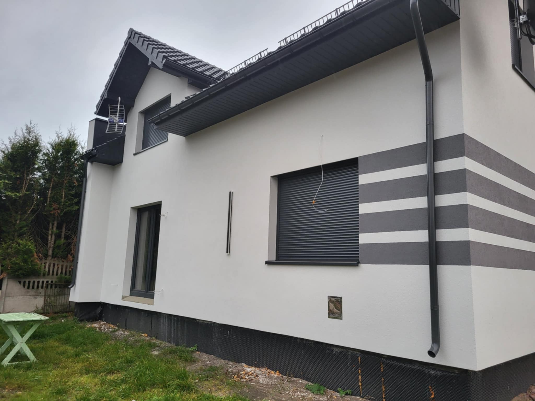 Boczny widok domu po renowacji elewacji: białe ściany z szarymi pasami, czarne okna i rynny. Widoczna antena na dachu i zielony stolik w ogrodzie.