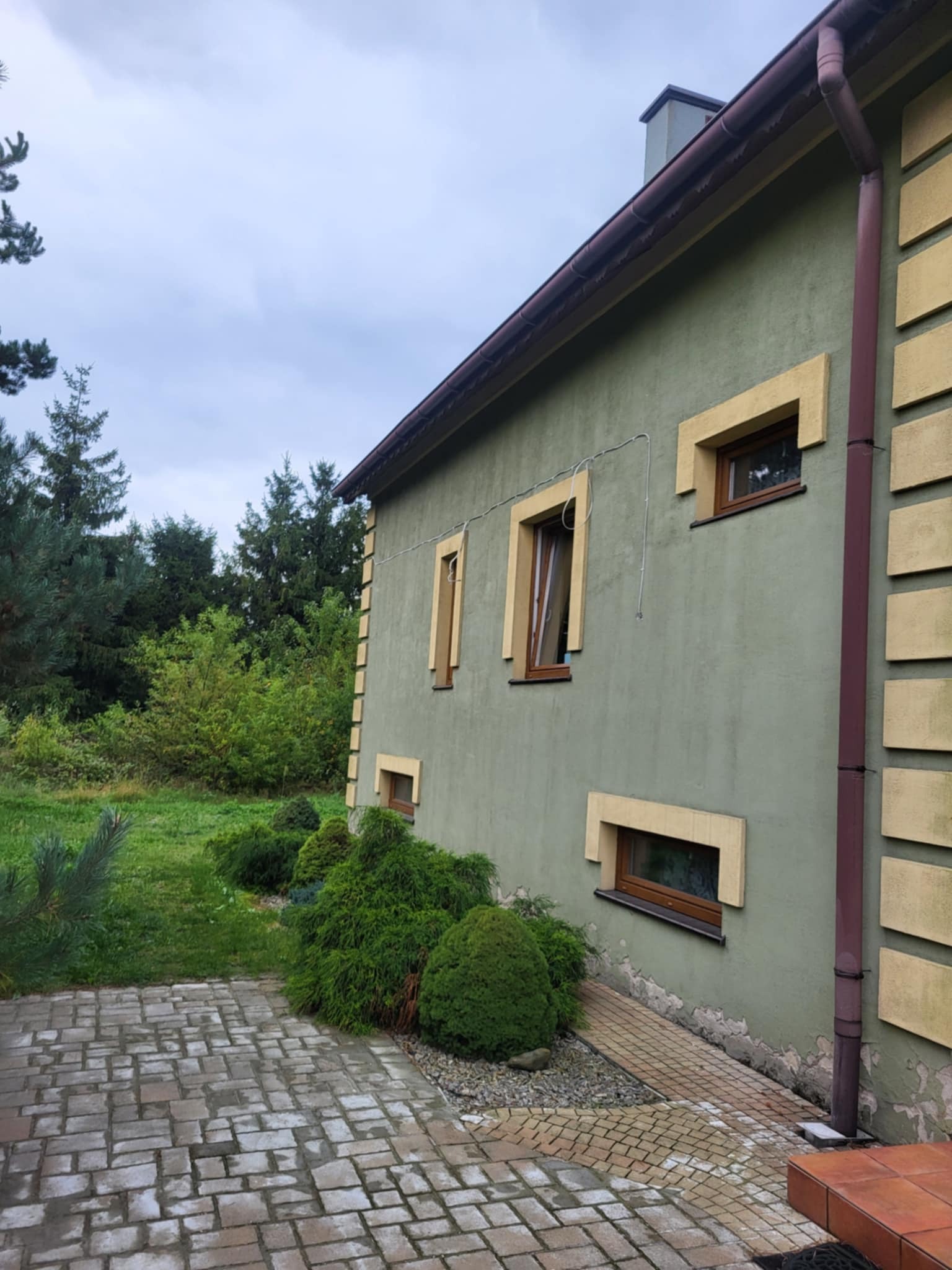 Elewacja domu z oknami w żółtych obramowaniach, widok z boku, w Trzyciążu. Widoczne krzewy i chodnik z kostki brukowej. Ściana w kolorze oliwkowym.