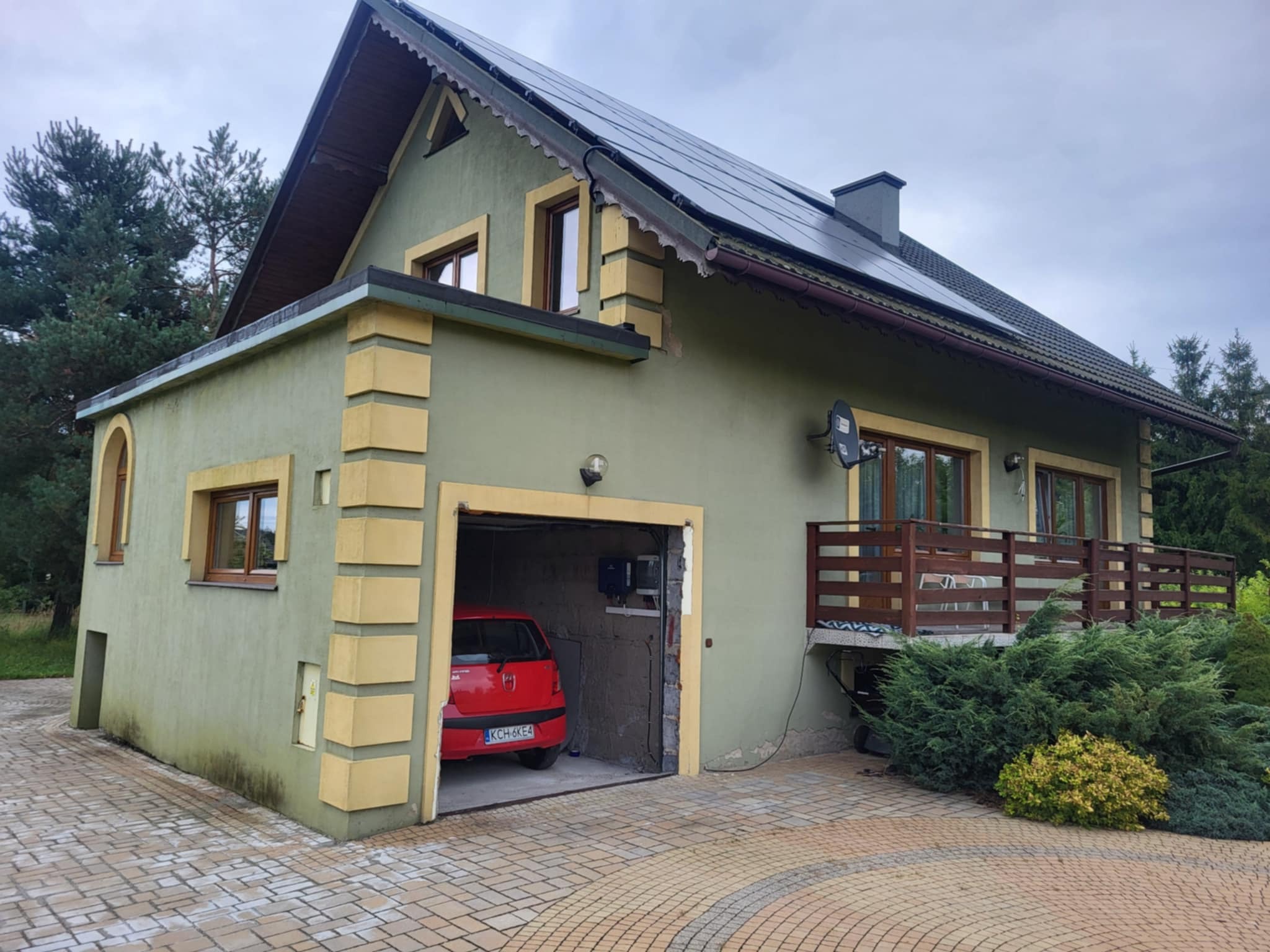 Dom jednorodzinny z panelami słonecznymi na dachu, garażem z czerwonym autem i balkonem z drewnianą balustradą. Elewacja w odcieniach zieleni z żółtymi detalami.