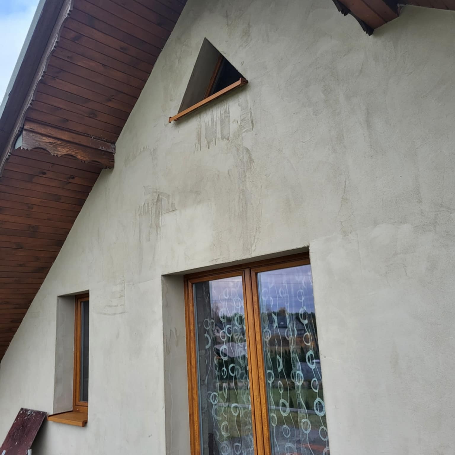 Szara elewacja domu jednorodzinnego z drewnianą stolarką okienną i trójkątnym oknem na poddaszu. Widoczne ślady po pracach wykończeniowych.