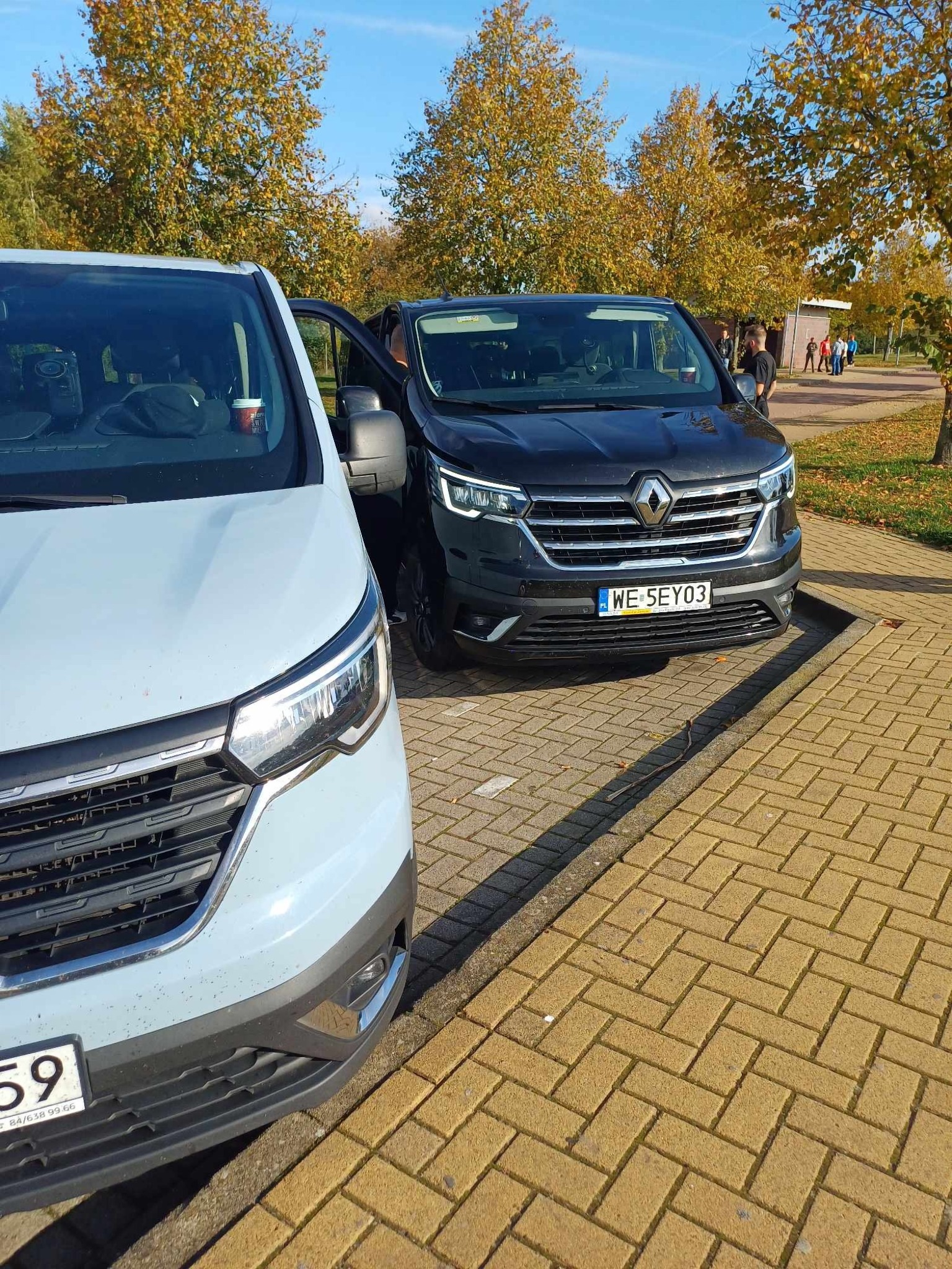 Dwa busy Renault, jeden jasnoniebieski i drugi ciemnoszary, zaparkowane na chodniku z kostki brukowej w jesienny dzień, z otwartymi drzwiami w ciemnoszarym busie i kubkiem z napojem...