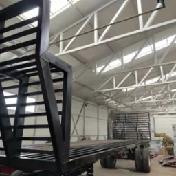 Czarna, metalowa konstrukcja przyczepy z drabinkową zabudową, widoczna w hali o białej konstrukcji dachu. Ujęcie z dołu podkreśla solidność wykonania.