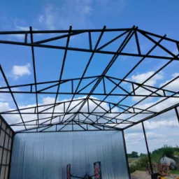 Konstrukcja stalowa hali w trakcie budowy, widok od dołu na metalowy szkielet i fragment obudowy, błękitne niebo w tle.