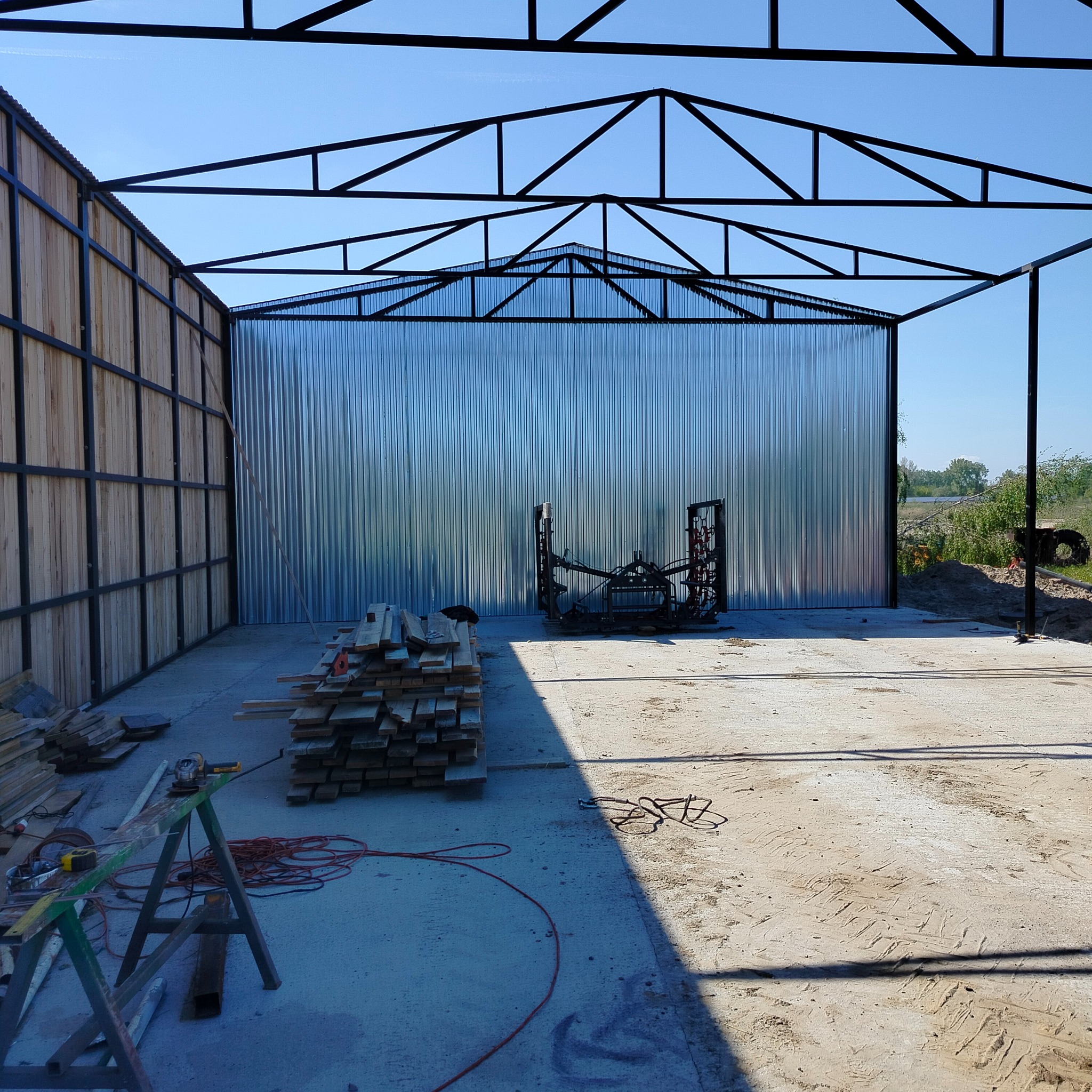 Wnętrze hali w budowie z metalową konstrukcją dachu i ścianami z blachy falistej. Na podłodze leżą deski i narzędzia, co sugeruje trwające prace wykończeniowe.