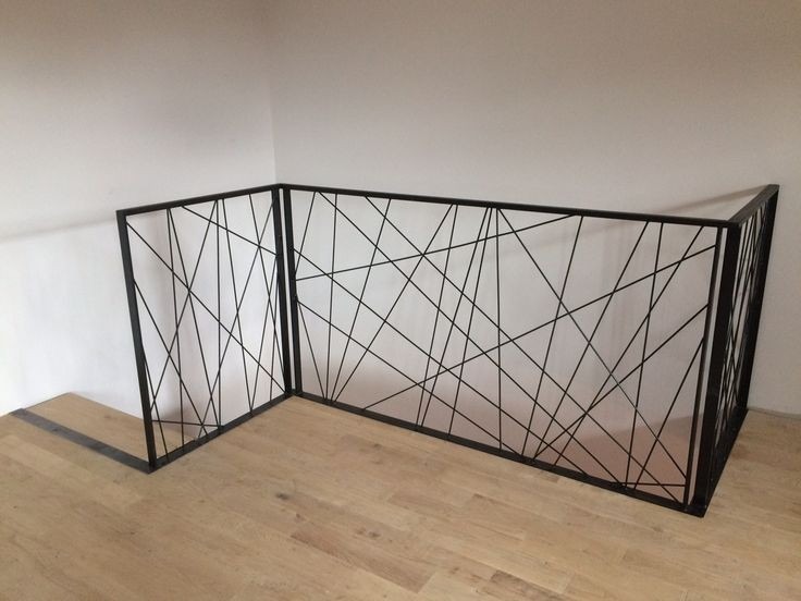 Czarna, metalowa balustrada schodowa o geometrycznym wzorze z cienkich prętów, zamontowana w rogu pomieszczenia z drewnianą podłogą i białymi ścianami.