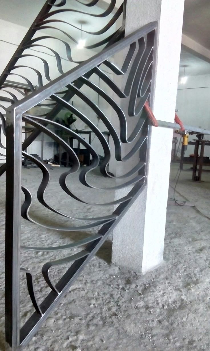 Metalowa konstrukcja schodowa z dekoracyjnym wypełnieniem, przymocowana do betonowego słupa za pomocą zacisku. Widoczne narzędzia w tle.