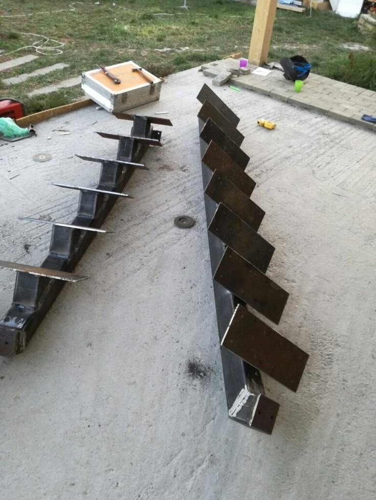 Dwa metalowe elementy konstrukcyjne schodów z przyspawanymi stopniami, leżące na szarym betonie. Widoczne ślady spawania i narzędzia w tle.