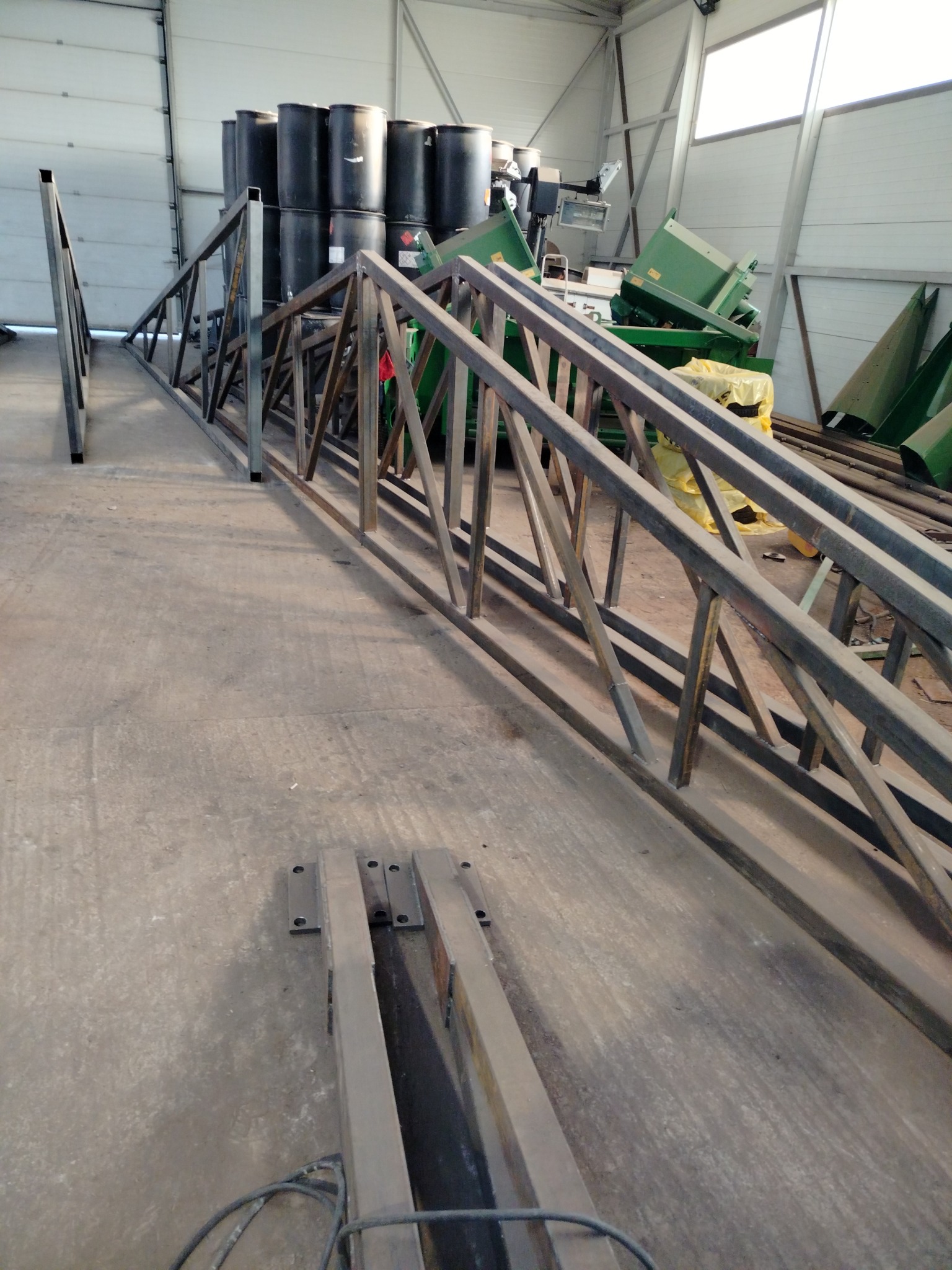 Elementy konstrukcji stalowej w hali produkcyjnej, widoczne spawane łączenia, w tle czarne beczki i maszyny. Surowy, przemysłowy charakter.