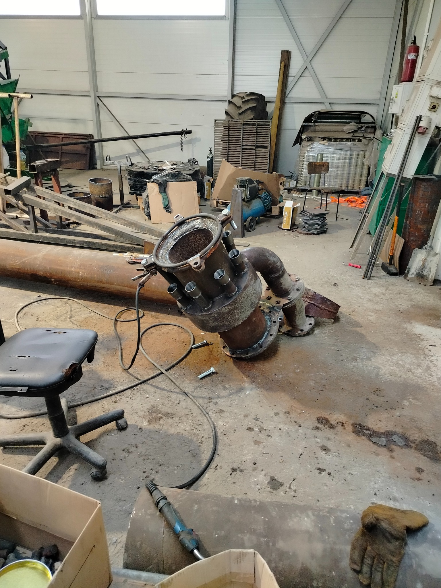 Metalowa konstrukcja rurowa z licznymi otworami, leżąca na podłodze warsztatu, obok krzesła i narzędzi. Widoczne spawy i ślady użytkowania.