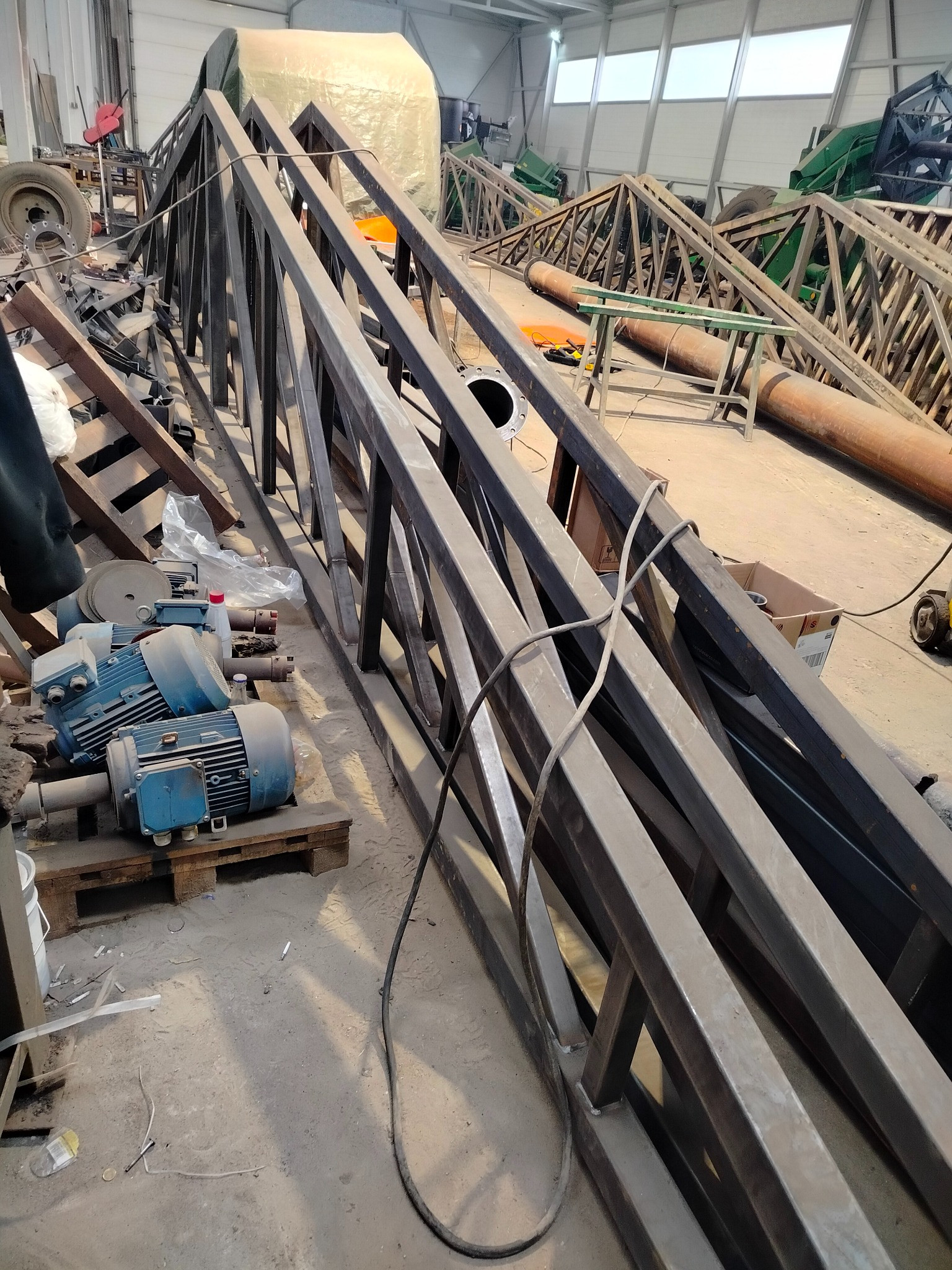 Elementy konstrukcji stalowej hali w warsztacie, z widocznymi spawami i silnikami elektrycznymi. W tle inne elementy i narzędzia.