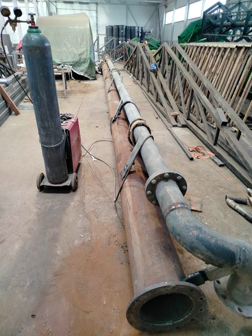 Długa, spawana rura z kołnierzami na tle hali, obok butla z gazem i spawarka. Widoczne metalowe konstrukcje w tle. Rura leży na innej, większej rurze.