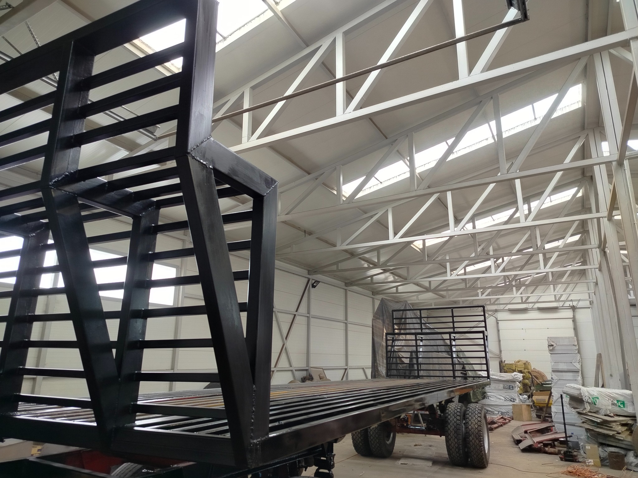 Czarna, metalowa konstrukcja przyczepy z drabinkową zabudową, widoczna w hali o białej konstrukcji dachu. Ujęcie z dołu podkreśla solidność wykonania.