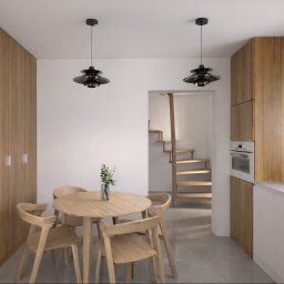 Jasne wnętrze: drewniane szafy, okrągły stół z krzesłami, czarne lampy wiszące. W tle schody. Minimalistyczny design z naturalnymi materiałami i subtelnym oświetleniem.