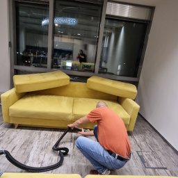 FORMICA Sp. z o.o. - Mężczyzna w pomarańczowej koszulce klęczy na podłodze, czyszcząc żółtą kanapę za pomocą odkurzacza piorącego; widoczne duże okna biurowe w tle.