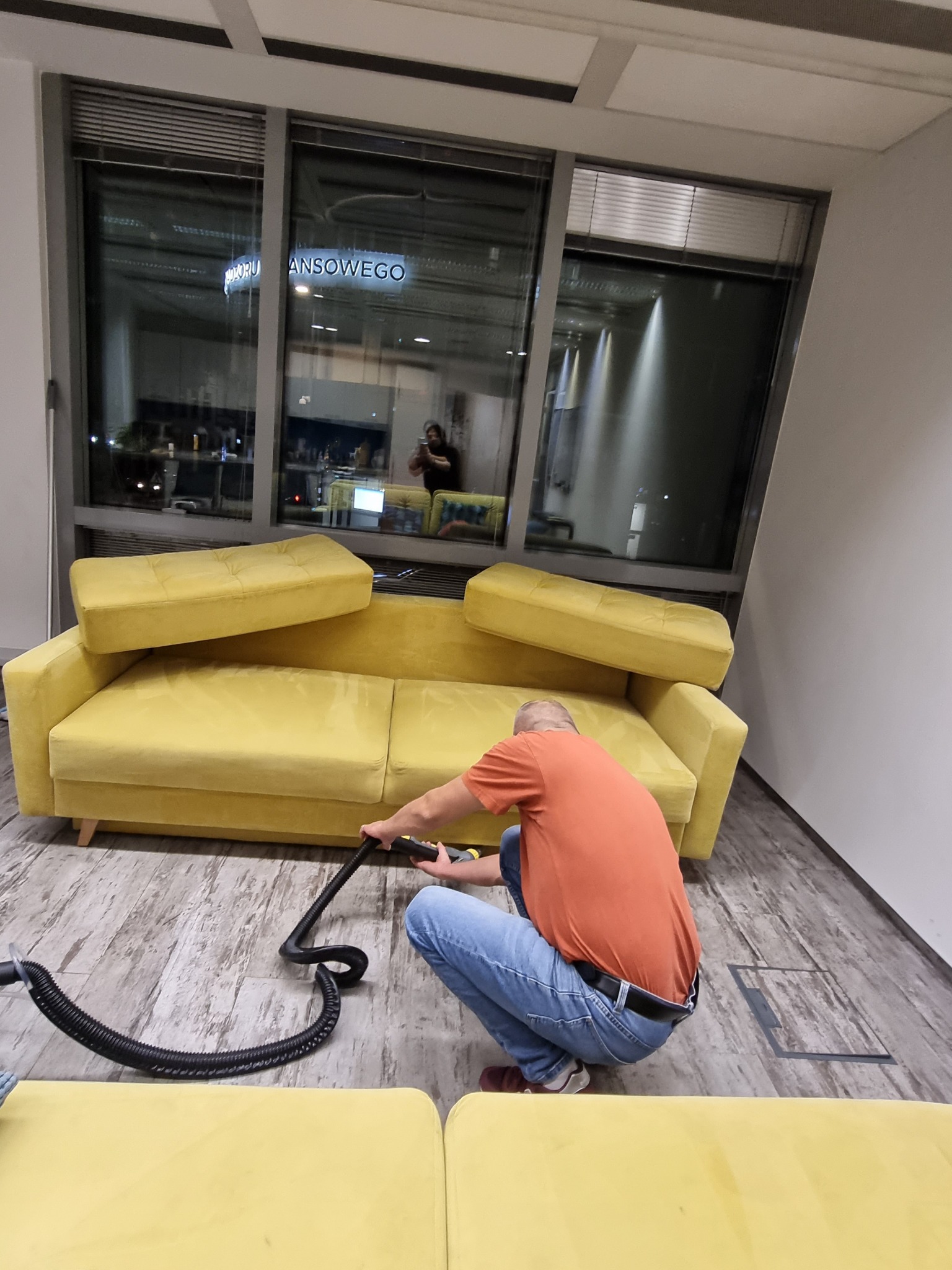 Mężczyzna w pomarańczowej koszulce klęczy na podłodze, czyszcząc żółtą kanapę za pomocą odkurzacza piorącego; widoczne duże okna biurowe w tle.