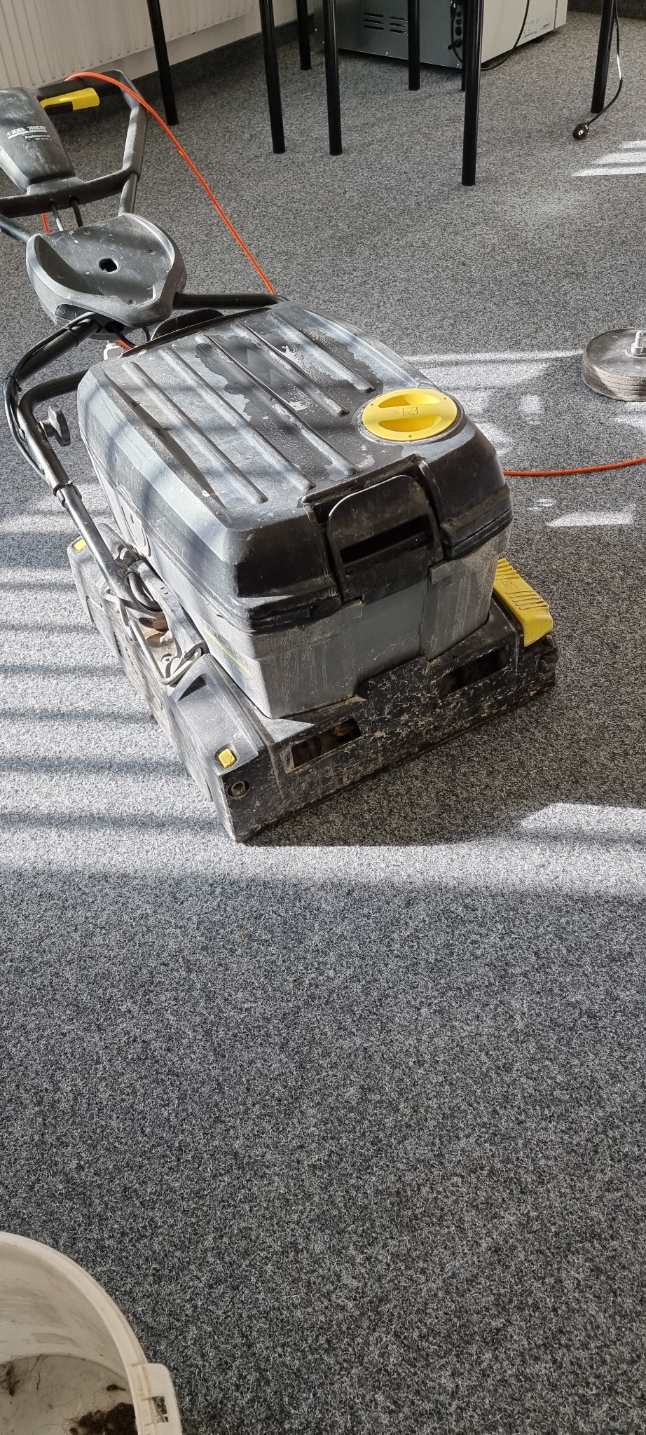 Profesjonalny automat do czyszczenia wykładzin dywanowych, widoczne ślady użytkowania, na szarym dywanie w biurze, z widocznymi promieniami słonecznymi.