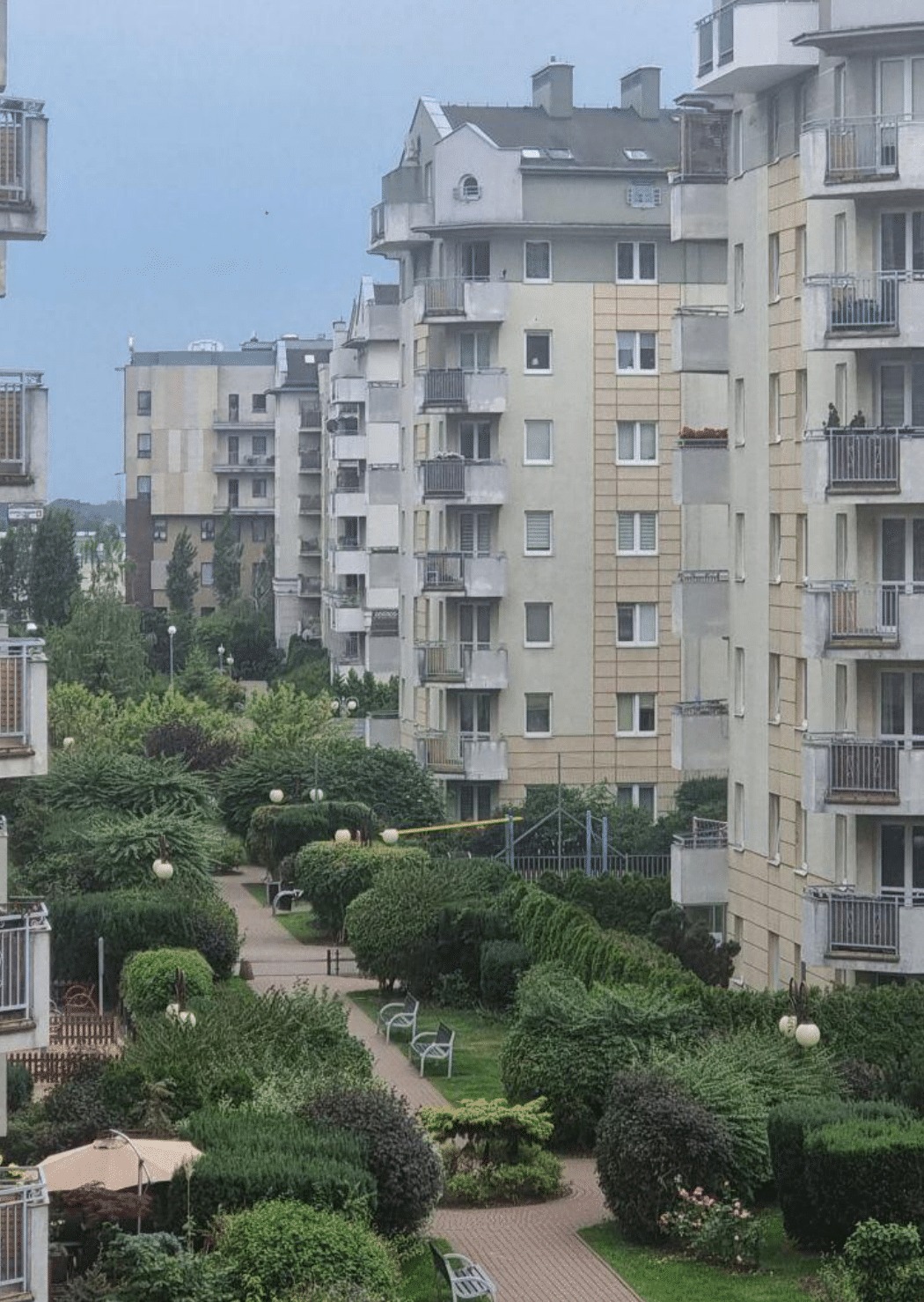 Widok z góry na zadbane osiedle mieszkaniowe z licznymi krzewami, drzewami i alejkami spacerowymi pomiędzy budynkami.