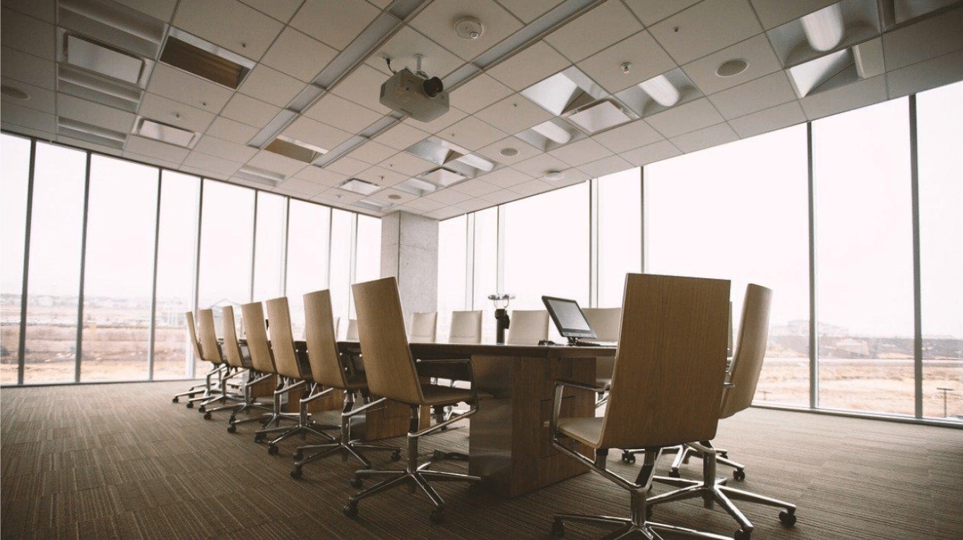 Jasna sala konferencyjna z dużym oknem, długim stołem i krzesłami na kółkach, projektor zamontowany na suficie i laptop na stole.