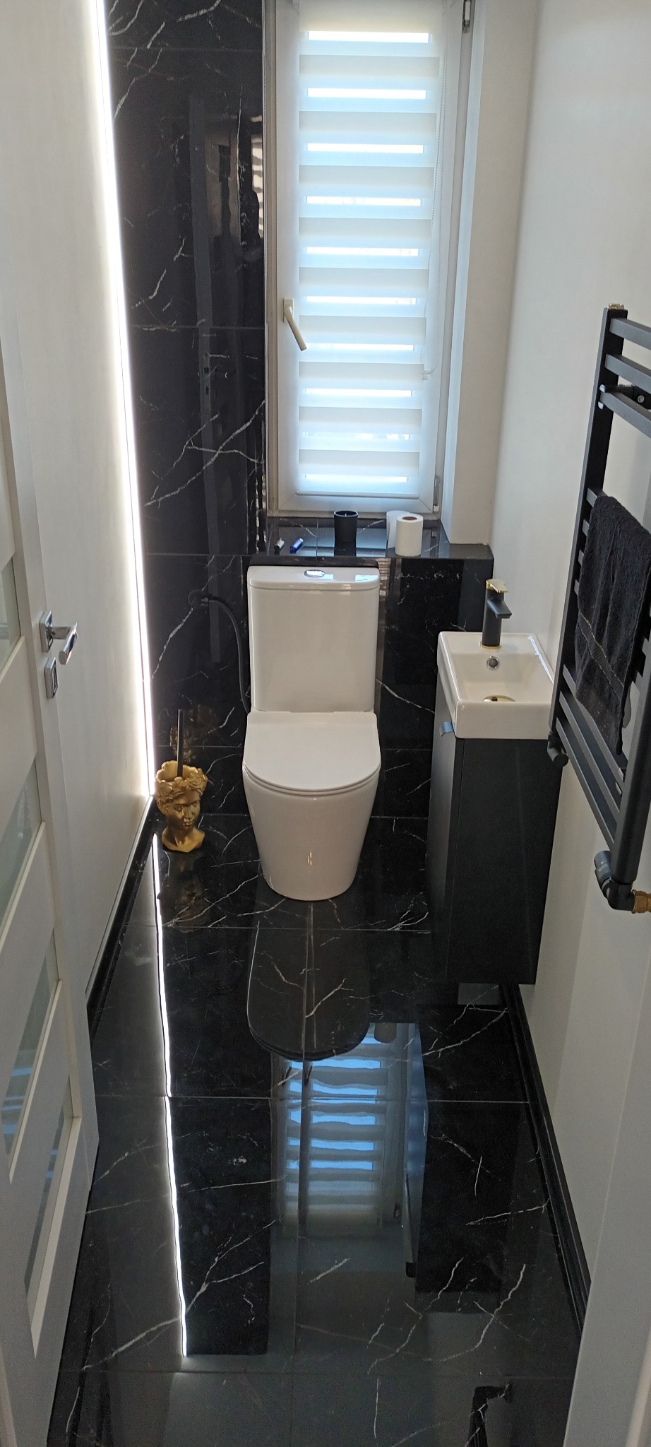 Wykończona łazienka z czarnymi marmurowymi płytkami podłogowymi i ściennymi, białą toaletą, małą czarną umywalką z złotą baterią i grzejnikiem drabinkowym z czarnym ręcznikiem.