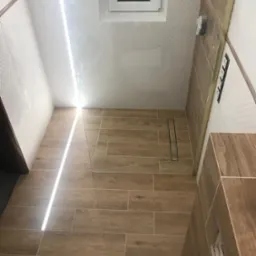 Wykończona łazienka z drewnopodobnymi płytkami, kabiną prysznicową ze szklaną ścianką i odpływem liniowym, widok z góry, częściowo widoczna toaleta.