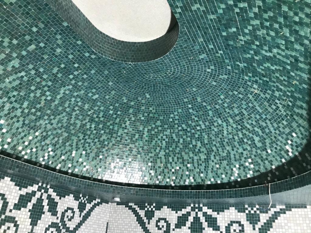 Mozaika z drobnych, zielonych płytek ceramicznych tworząca półokrągłą powierzchnię, z widocznym fragmentem białej mozaiki w tle i dekoracyjnym wzorem na obrzeżu.