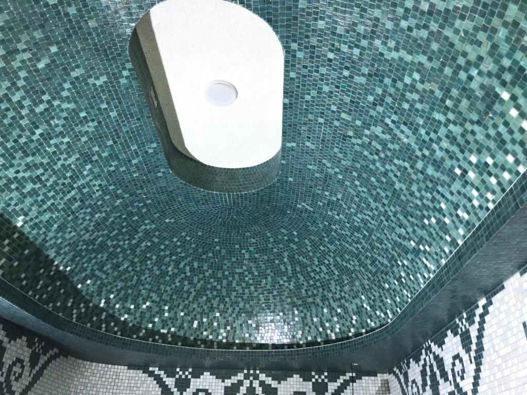 Mozaika z drobnych, turkusowych płytek na zaokrąglonym suficie, z centralnie umieszczonym, białym oświetleniem; dolna część ściany wykończona mozaiką z motywem geometrycznym w odcieniach szarości...