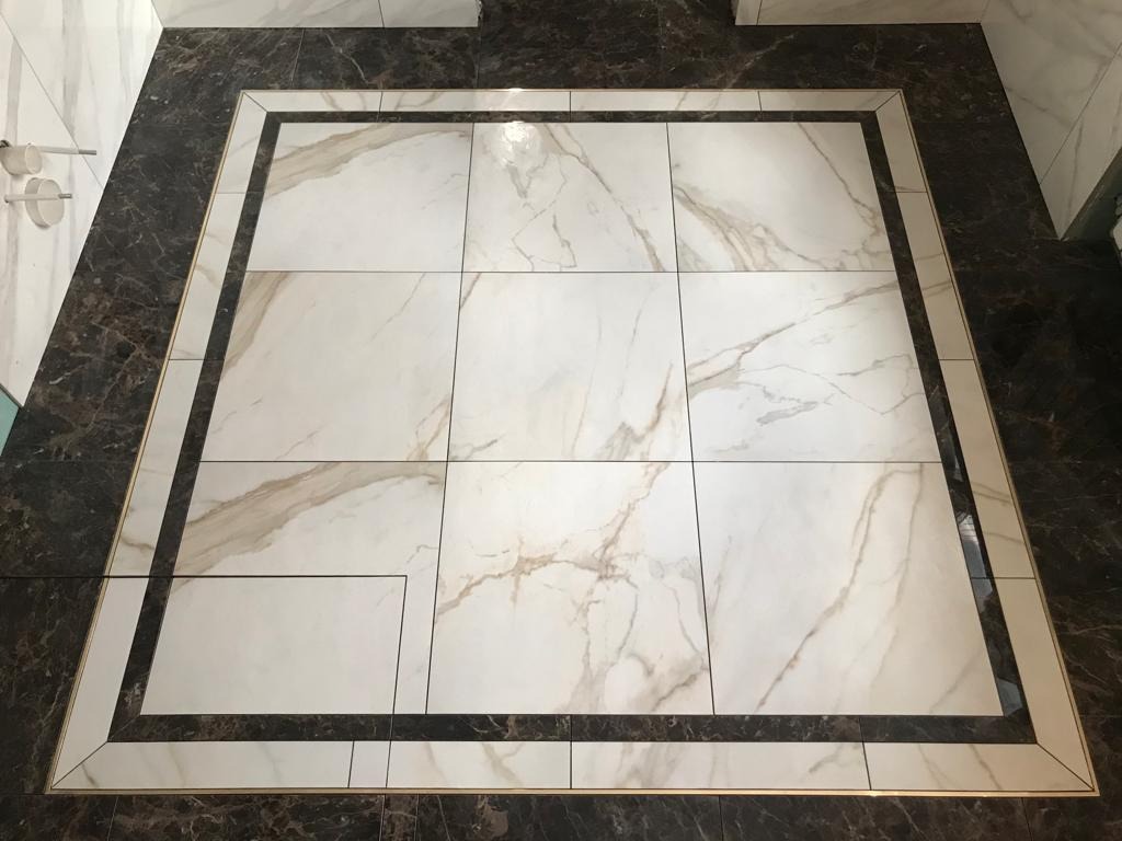 Wykończona podłoga z białych płytek imitujących marmur, obramowana ciemnymi płytkami i złotą listwą dekoracyjną.