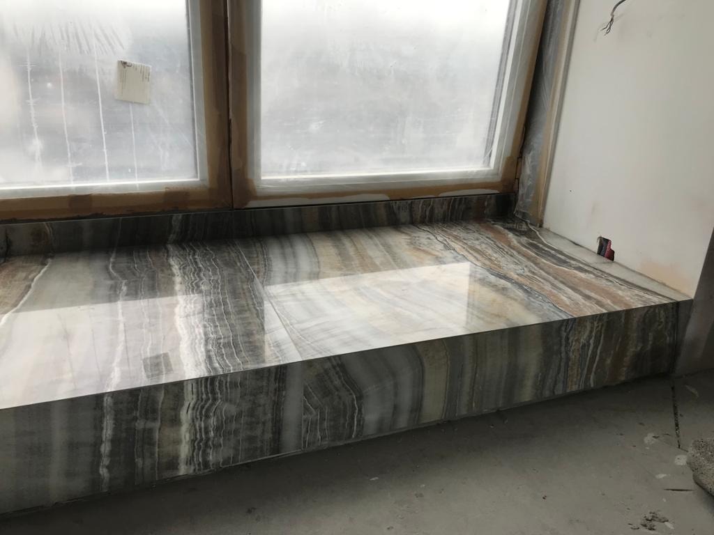 Parapet okienny wyłożony polerowanym kamieniem o wyraźnym, warstwowym wzorze w odcieniach szarości, brązu i bieli, widoczne niedoskonałości ściany i okna sugerują trwający remont.