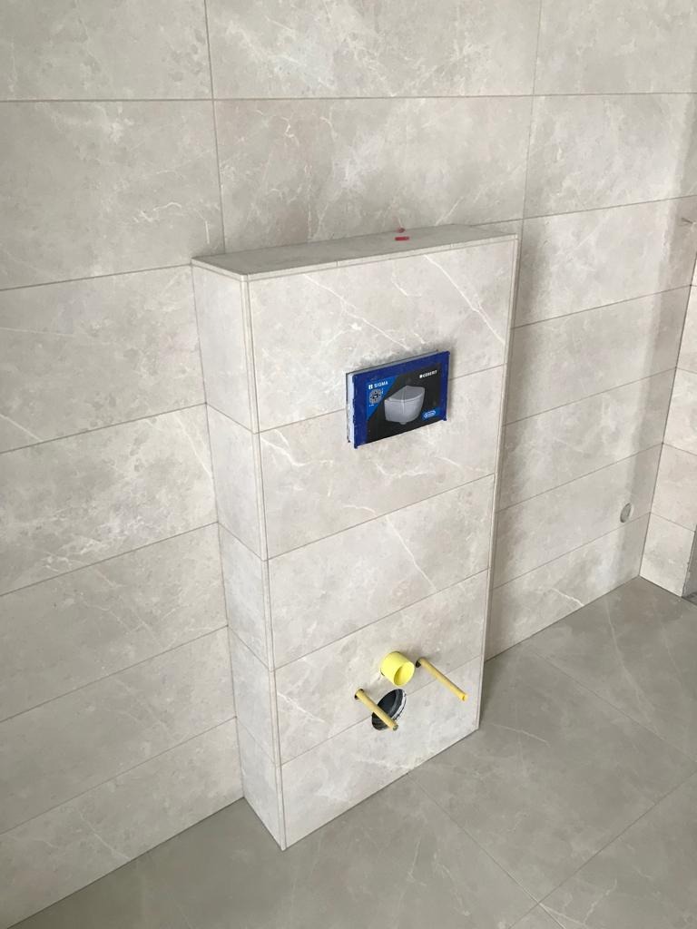 Obudowa stelaża podtynkowego WC wykończona płytkami imitującymi marmur, dopasowanymi do płytek ściennych i podłogowych. Widoczne żółte zaślepki instalacyjne.