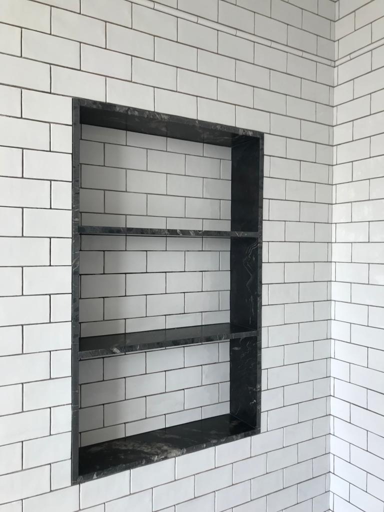 Wnęka z półkami wykończona czarnym marmurem w ścianie z białych, prostokątnych płytek ułożonych w cegiełkę.
