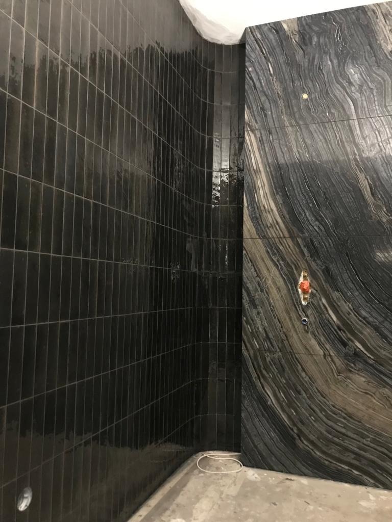 Ciemne, błyszczące, pionowo ułożone płytki w wąskim formacie na zakrzywionej ścianie kabiny prysznicowej, sąsiadujące z płytą z naturalnego kamienia o wyraźnym, falistym wzorze i zamontowanym...