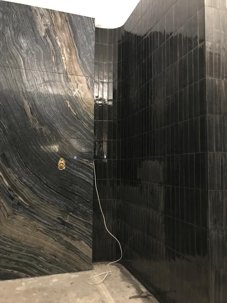 Nowoczesna łazienka z kontrastującymi płytkami: ściana z płytek imitujących marmur z widoczną armaturą i ściana wyłożona pionowymi, czarnymi płytkami. Na podłodze widoczne ślady zaprawy.