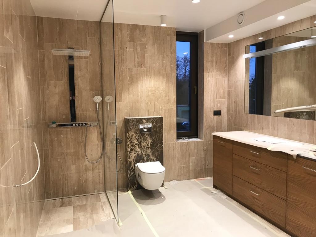 Nowoczesna łazienka z beżowymi płytkami imitującymi trawertyn, kabiną prysznicową ze szkła, podwieszaną toaletą z marmurową obudową i drewnianą szafką pod umywalką. Na podłodze widoczne taśmy...