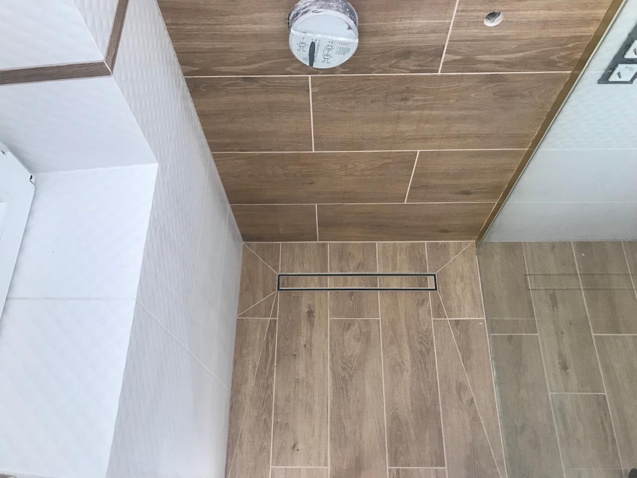 Wykończona łazienka z drewnopodobnymi płytkami na podłodze i suficie, z odpływem liniowym i białymi płytkami strukturalnymi na ścianie, widok z góry.