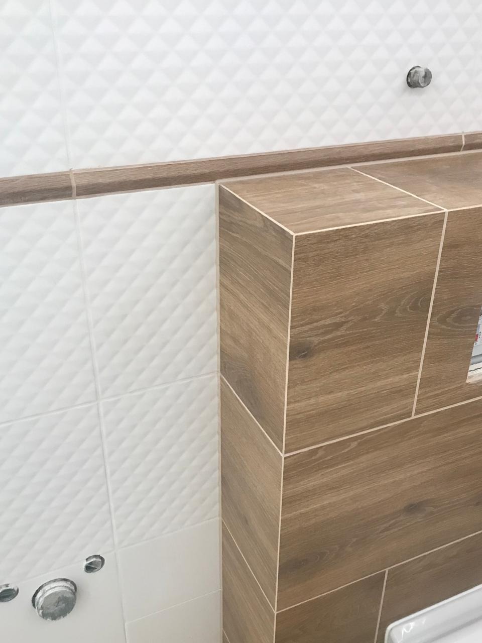 Detal wykończenia łazienki: białe, trójwymiarowe płytki ścienne kontrastują z drewnopodobną obudową stelaża WC, zakończoną listwą dekoracyjną.