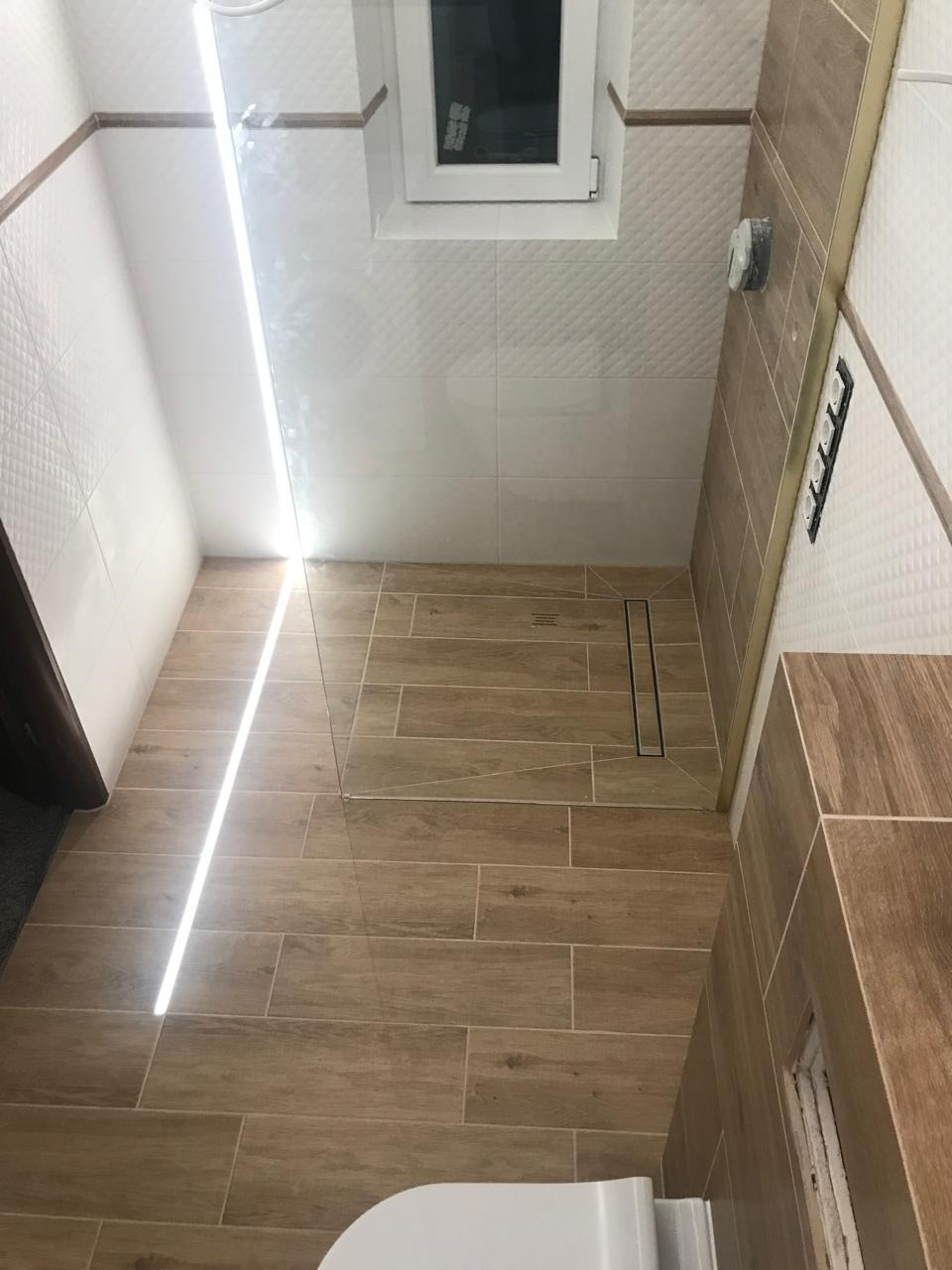 Wykończona łazienka z drewnopodobnymi płytkami, kabiną prysznicową ze szklaną ścianką i odpływem liniowym, widok z góry, częściowo widoczna toaleta.