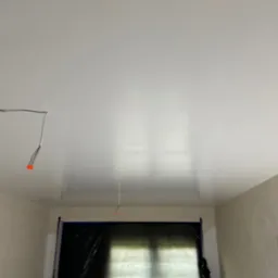 Wnętrze po remoncie: gładka, biała powierzchnia sufitu i ścian. Okno zabezpieczone folią malarską. Widoczne przewody elektryczne wystające z sufitu.