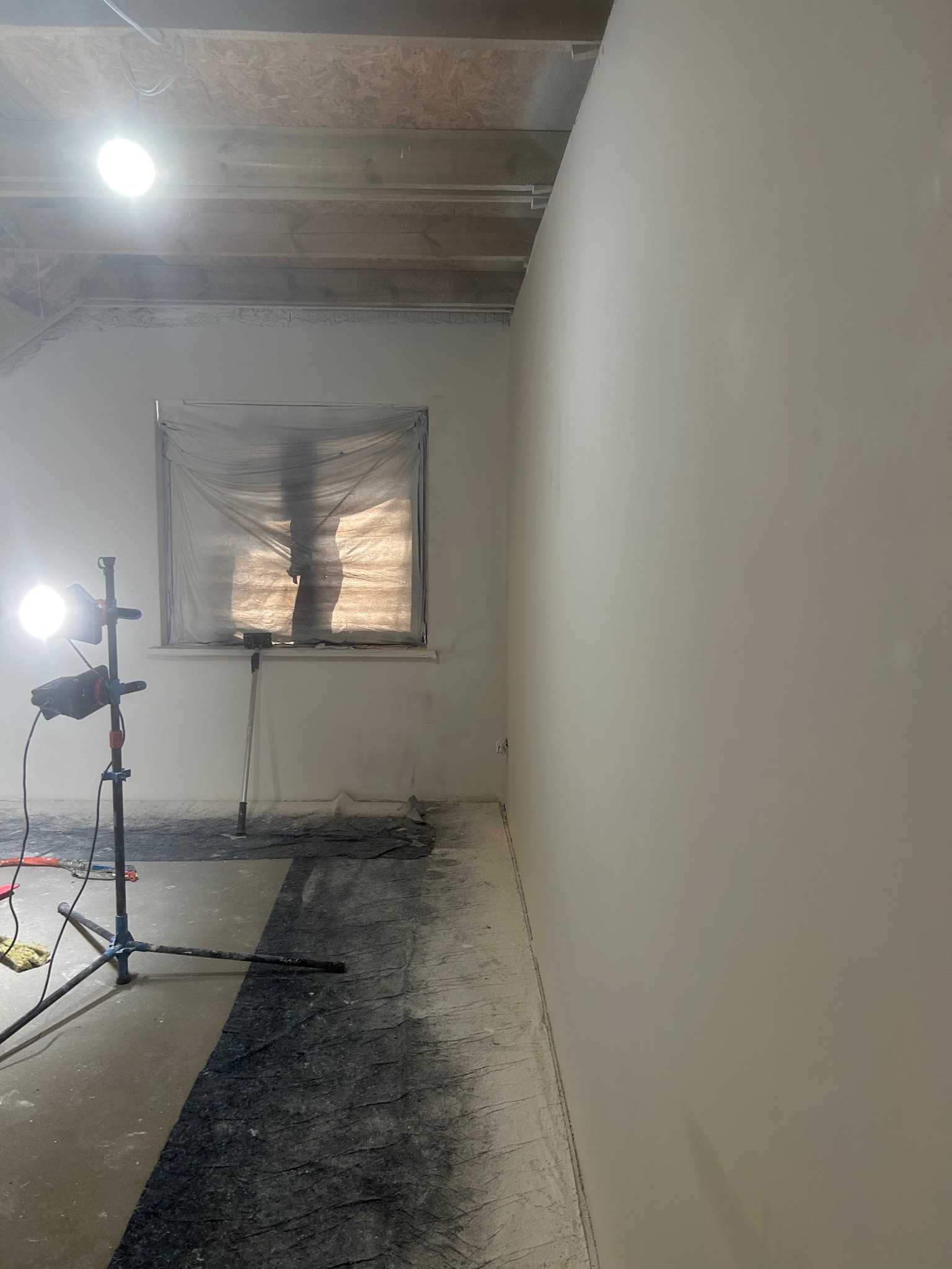 Wnętrze po szpachlowaniu: gładka, jasna ściana, okno zabezpieczone folią, lampa budowlana. Podłoga częściowo zakryta folią ochronną. Widoczne ślady pyłu po szpachlowaniu.