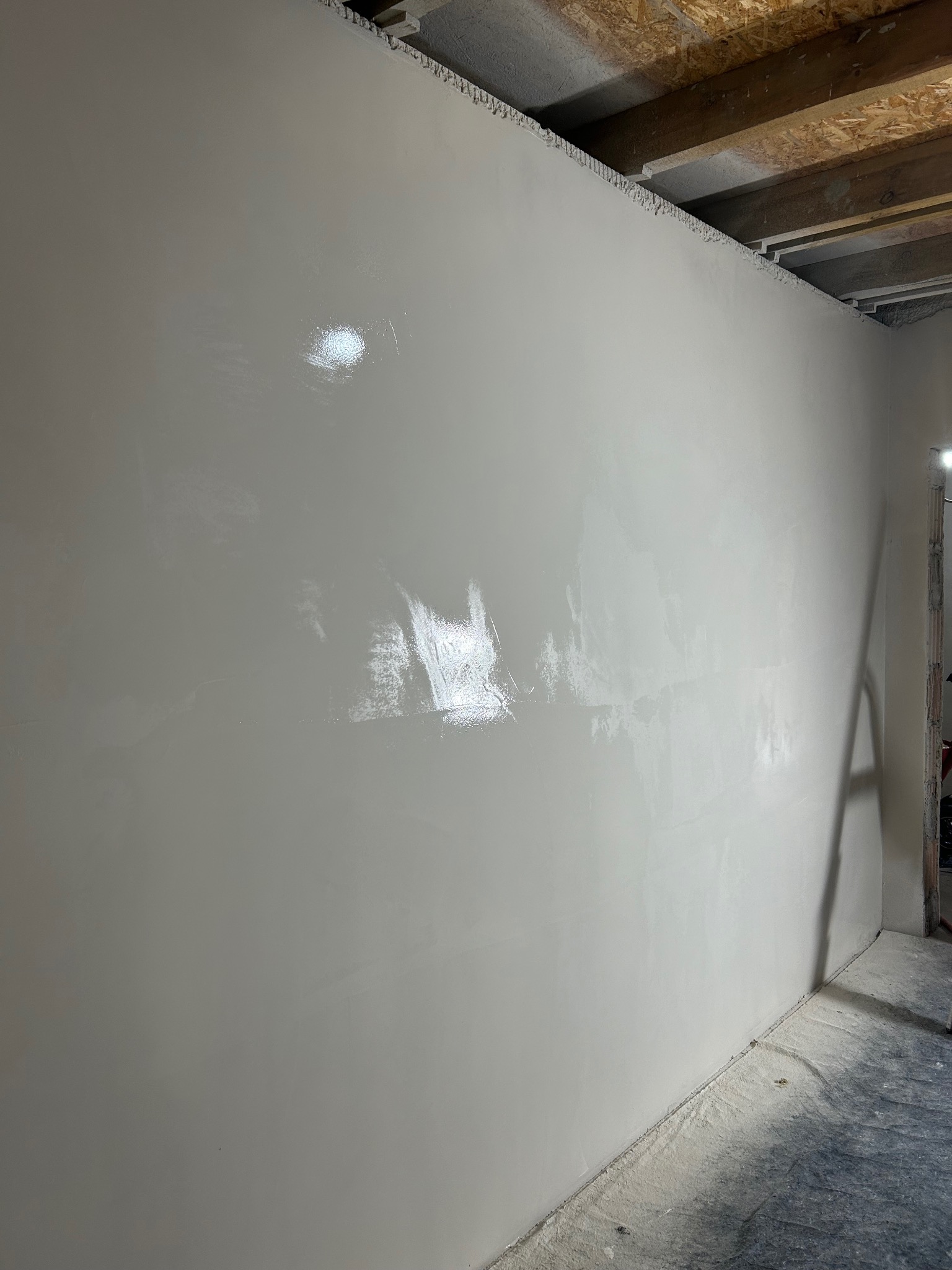 Ściana po nałożeniu gładzi, widoczne refleksy światła na gładkiej powierzchni. Surowy strop z drewnianych belek. Praca w trakcie wykończenia.