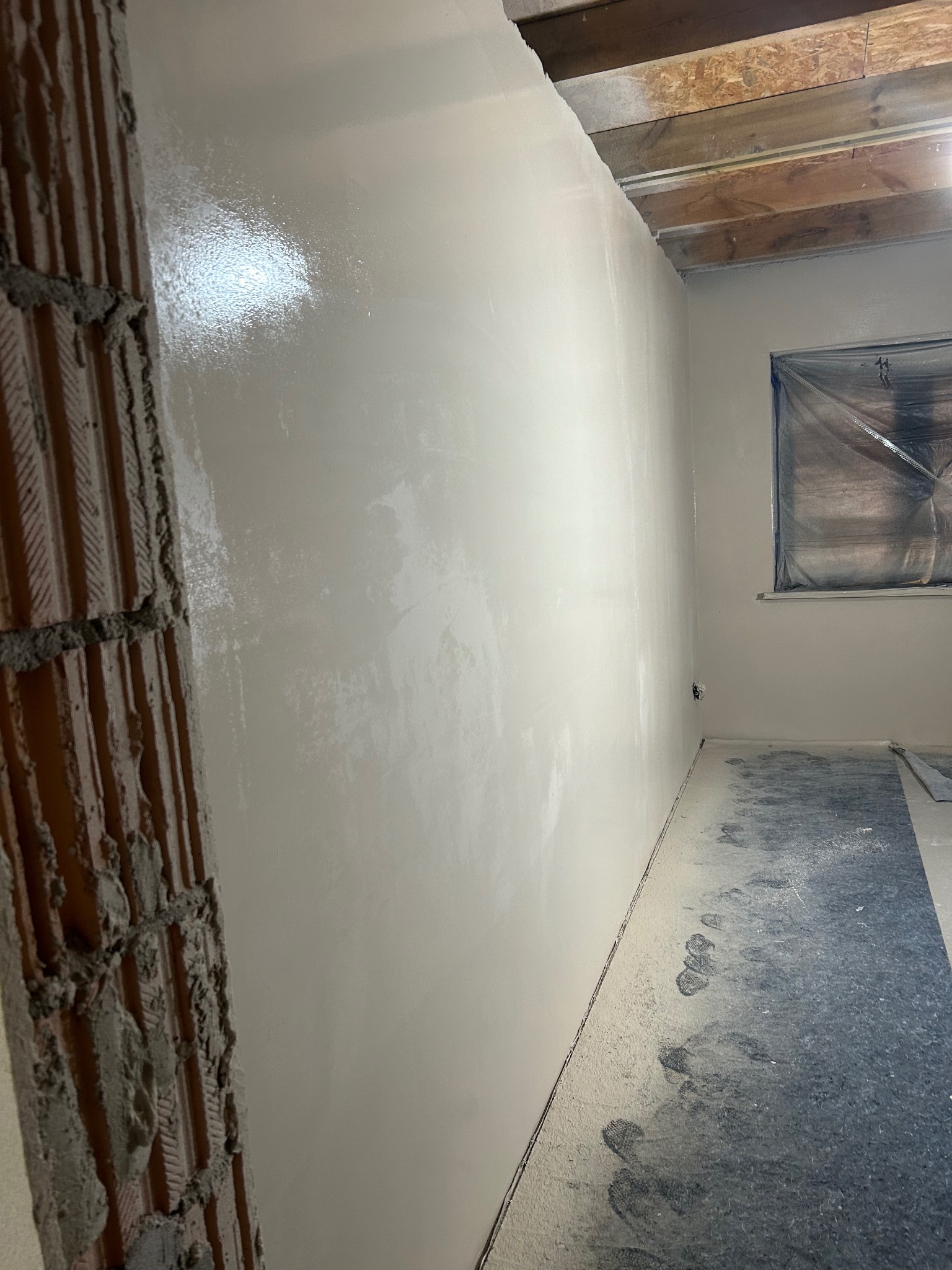 Świeżo położona, błyszcząca, jasna warstwa na ścianie w trakcie remontu. Widoczny fragment surowej cegły i okno zabezpieczone folią malarską.