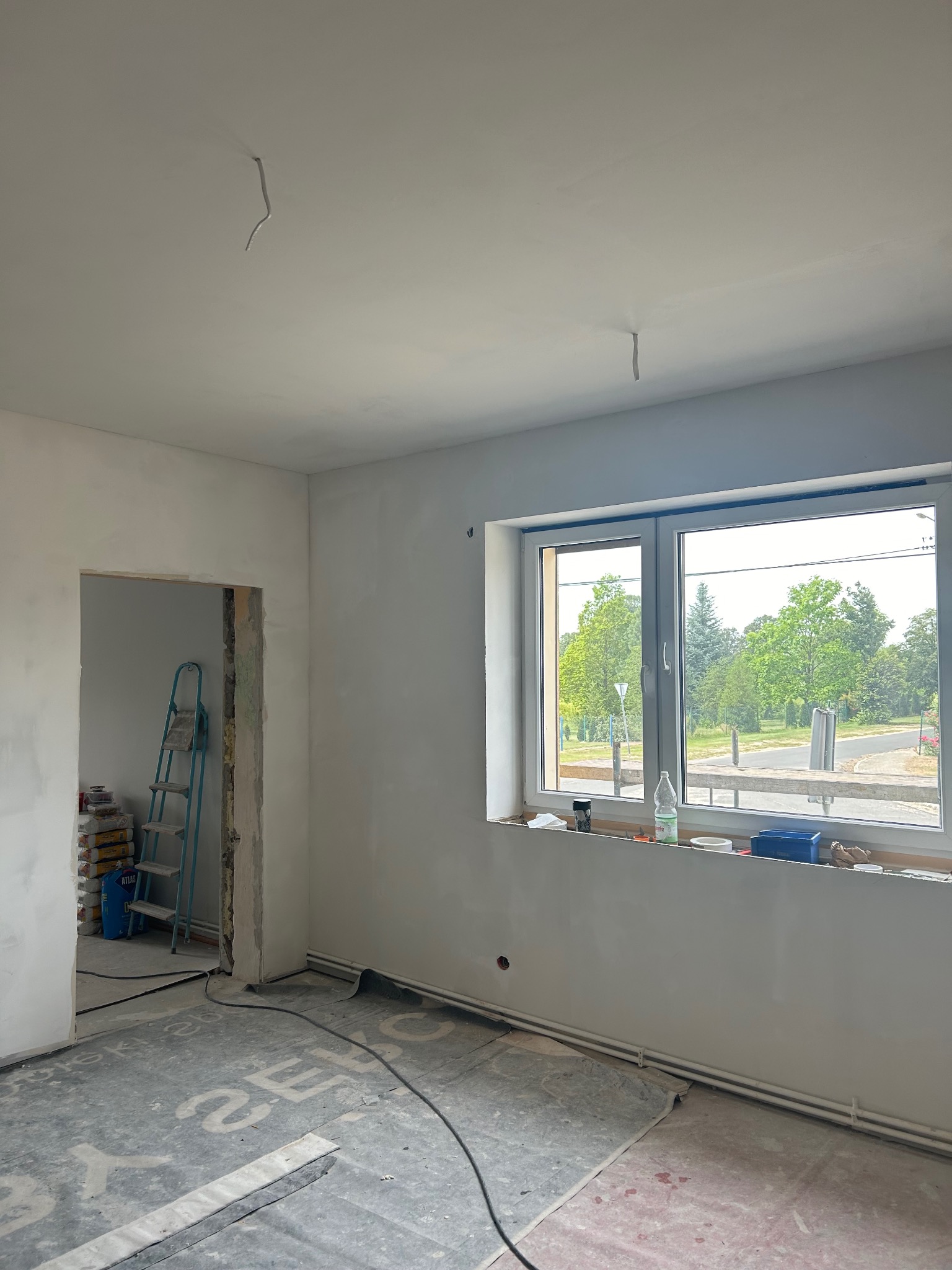 Wnętrze w trakcie remontu: ściany po wstępnym wyrównaniu, okno z widokiem na zieleń, drabina oparta o ścianę w przejściu. Podłoga zabezpieczona folią.