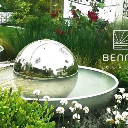 PRZEMYSŁAW BENNIG OGRODY - Nowoczesny ogród z kulistymi fontannami ze stali nierdzewnej, bujną roślinnością i białymi kamieniami. Logo firmy ogrodniczej w prawym górnym rogu.