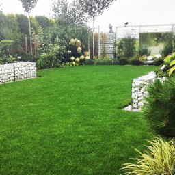 PRZEMYSŁAW BENNIG OGRODY - Zadbany trawnik z gabionami wypełnionymi białymi kamieniami, ozdobne krzewy i trawy. W tle białe ogrodzenie i srebrne kule. Logo firmy w prawym dolnym rogu.