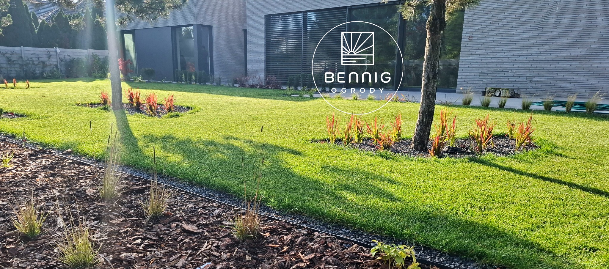 Nowoczesny ogród z równo przystrzyżonym trawnikiem, ozdobnymi trawami i korą. W tle minimalistyczny dom z dużymi oknami i logo firmy ogrodniczej.